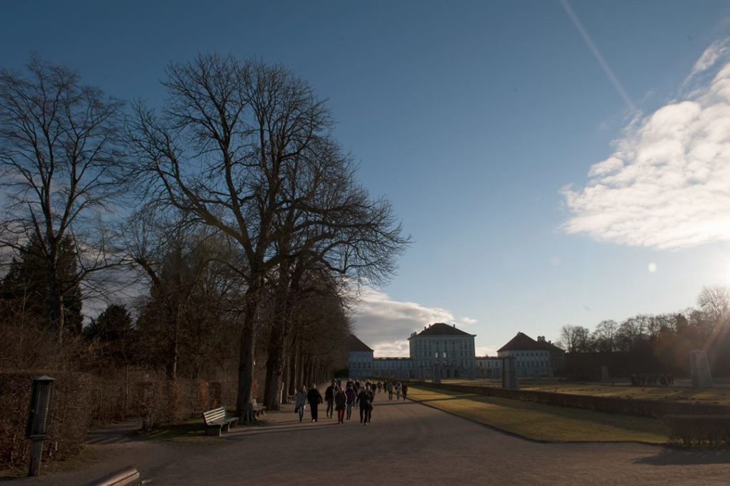 Eine Exkursionsgruppe geht im Schlosspark Nymphenburg beim Natura 2000-Gipfel in München durch den Schlosspark Nymphenburg auf einem Weg zwischen Baumreihe und Beeten. Im Hintergrund das Schloss Nymphenburg.