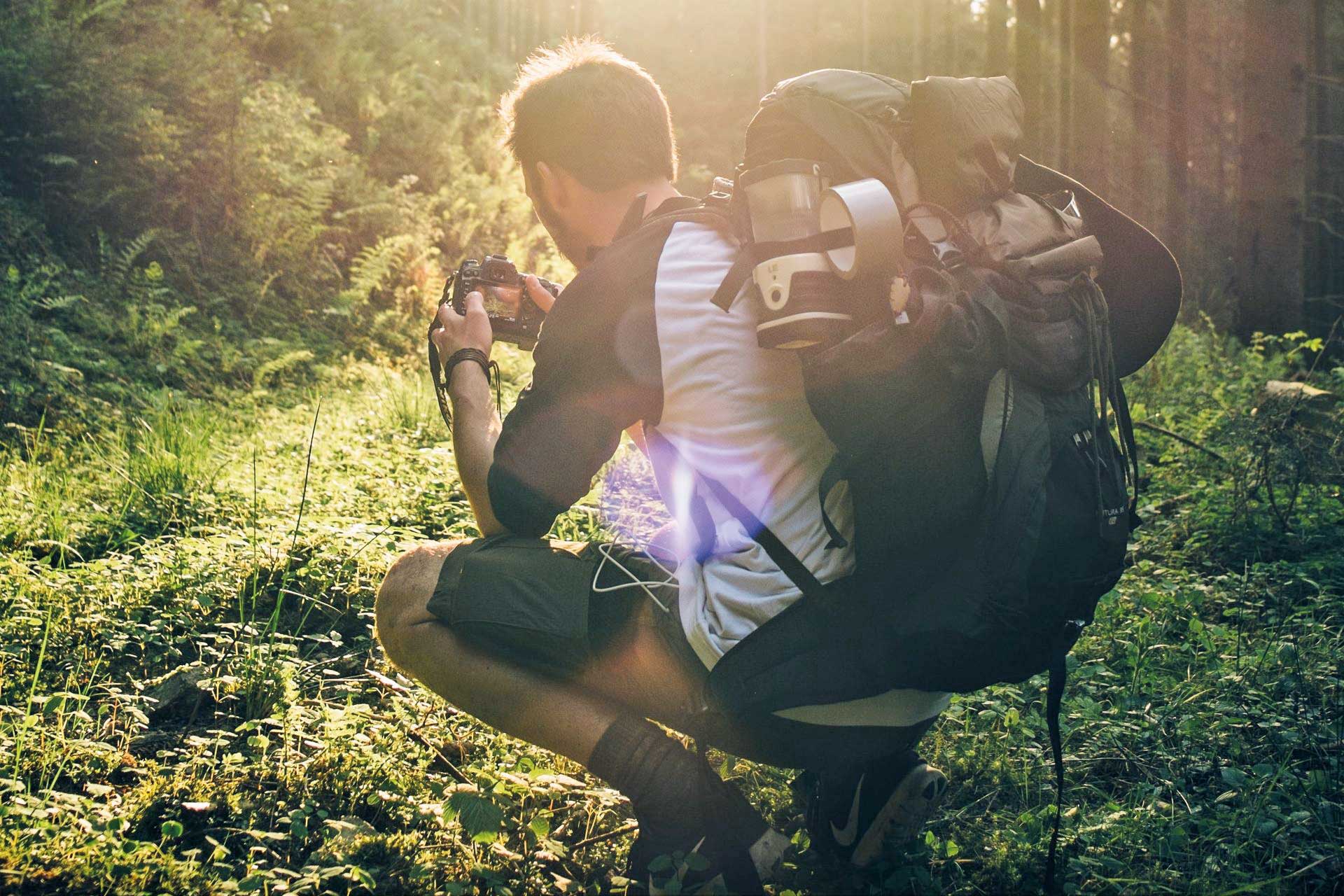 A man with a large hiking backpack crouches down to photograph the ground vegetation. He sits in a spruce forest, the sunlight catching his eye.