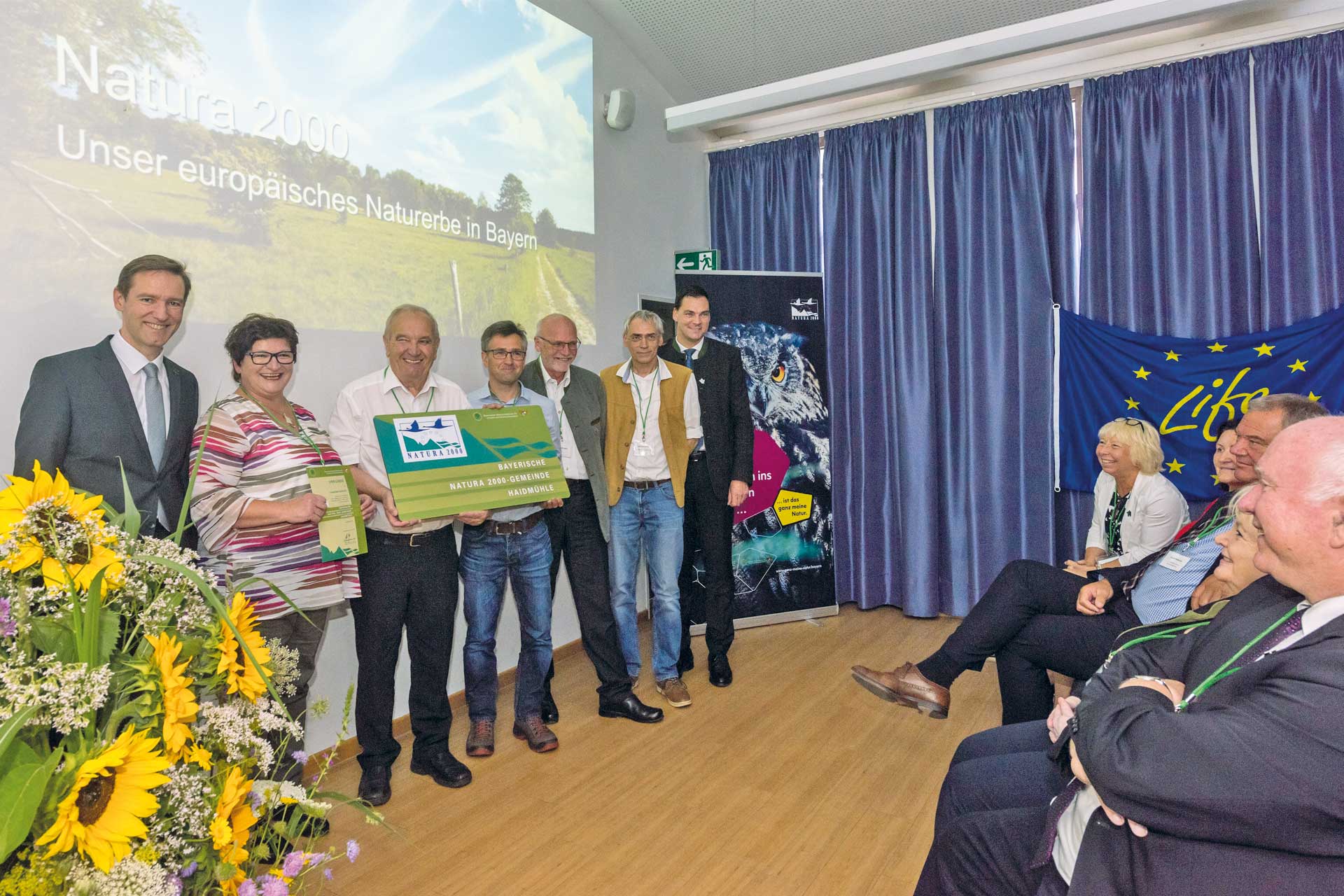 A middle-aged woman and four men stand in front of an audience, smiling into the camera. Two of the men hold a green sign reading 'Bavarian Natura 2000 Community of Haidmühle'