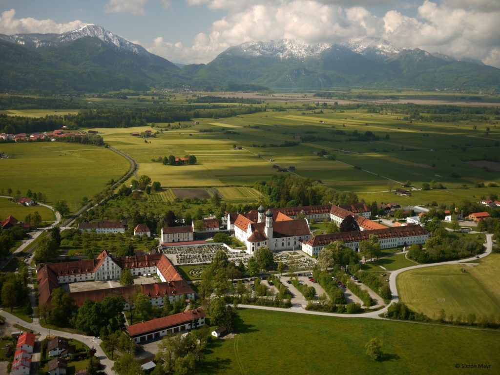 Luftbild einer Landschaft mit einem großen Kloster im Vordergrund, im Hintergrund Wiesen und Felder und Berge