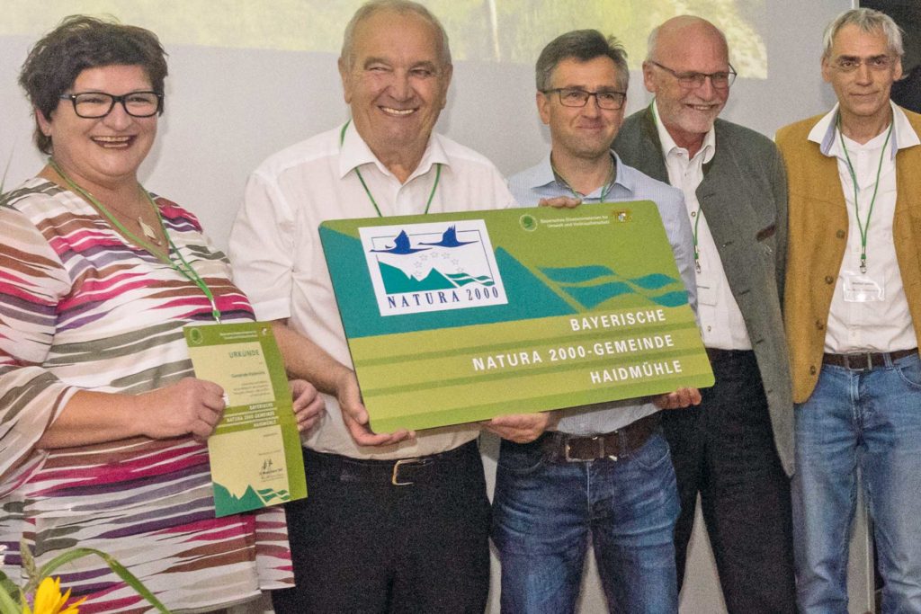 Eine Frau und vier Männer mittleren Alters schauen lächelnd in die Kamera. Zwei der Männer halten eine grüne Tafel mit der Aufschrift "Bayerische Natura 2000-Gemeinde Haidmühle".