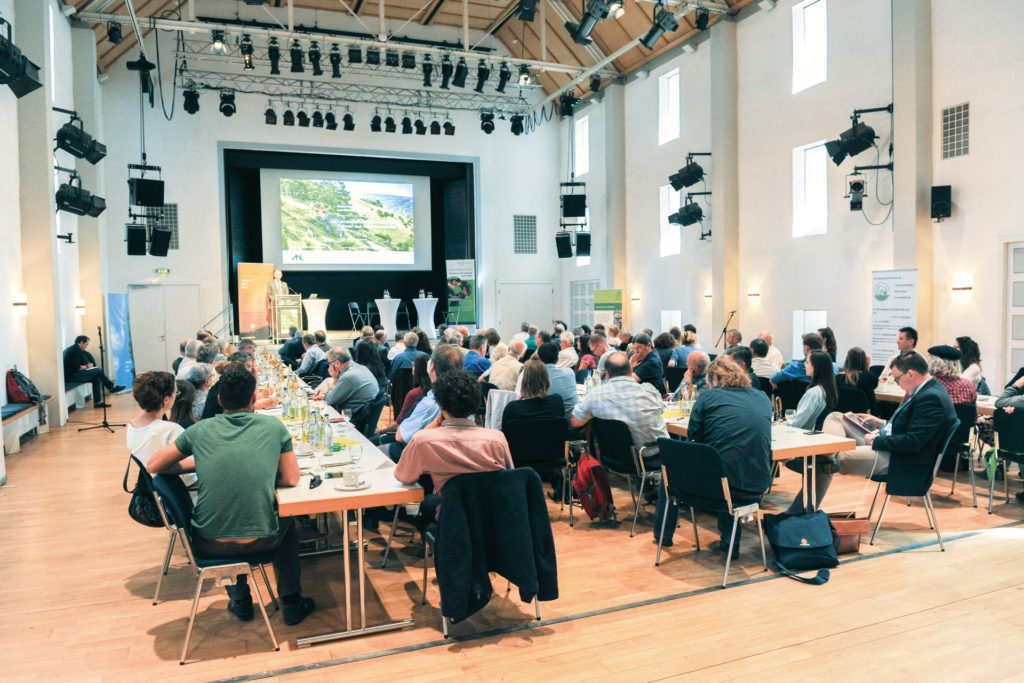 Menschen sitzen an langen Tischen in einem grßen Saal. Im Hintergrund steht ein Redner an einem Podium vor einer Leinwand.