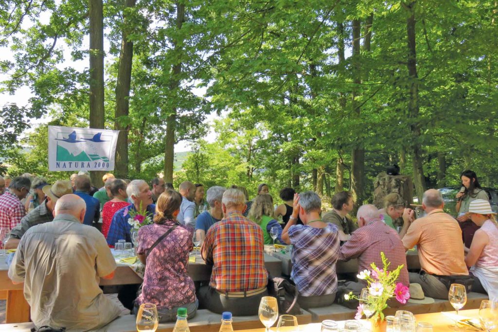 Gruppe von Menschen sitzen zusammen und verkosten Wein. Im Hintergrund weht eine Natura 2000 Fahne. Umgebung: Wald und Natur