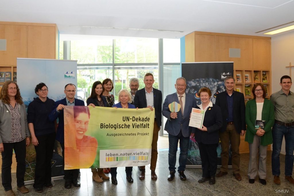 The LIFE living Natura 2000 team receives an award. The team and members of the ministry stand in front of campaign posters while holding a flag showing the award.