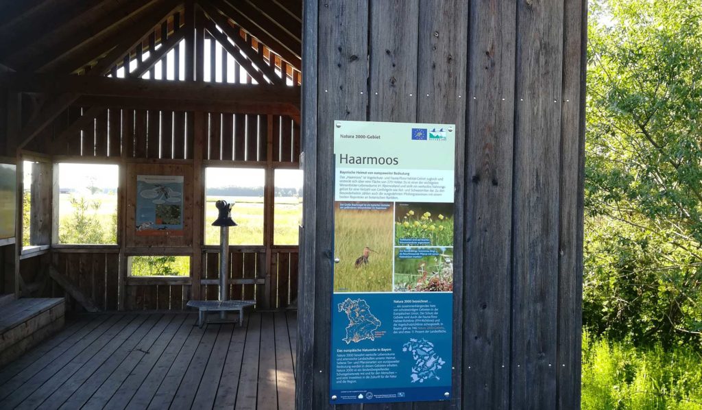 Blick auf eine Hütte im Freien mit Aussichtsglas im Inneren und mehreren Schildern mit Bildern und Texten zum Haarmoos.