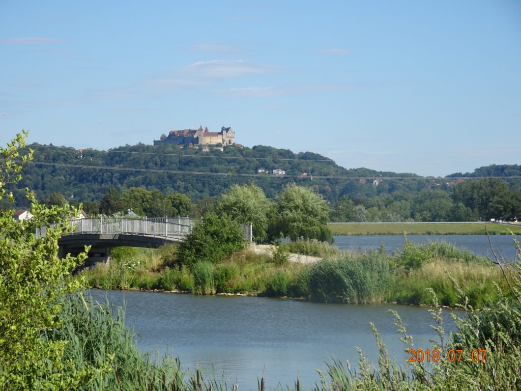 Auf dem Bild befindet sich im Vordergrund ein See, auch ist eine Straße mit einer Brücke zu sehen. Im Hintergrund befindet sich auf einem bewaldeten Hügelrücken eine Burg, die Veste Coburg.