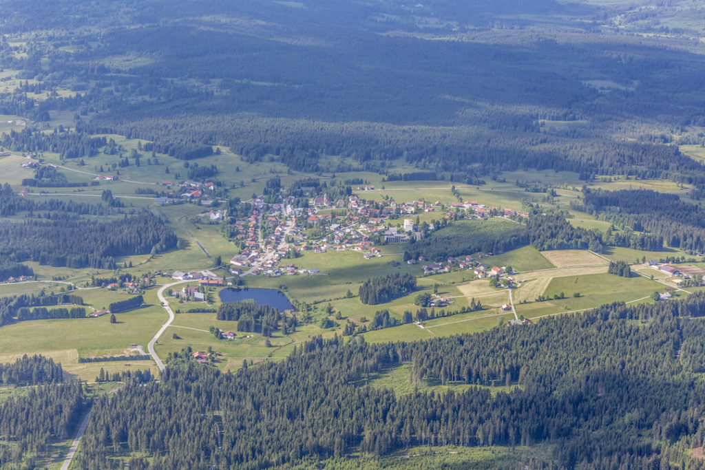 Eine Luftaufnahme der Gemeinde Haidmühle mit dem Dorf Haidmühle im Zentrum. Unmittelbar um das Dorf befinden sich grüne Weiden, Grünland und ein See. An die offenen Bereiche schließen sich weitläufige Fichtenwälder an.
