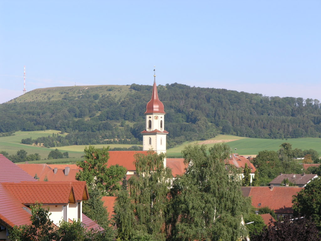 In der Bildmitte ist eine Kirche zu sehen, die Kirche von Röckingen. Im Bildhintergrund ist ein Berg, der Hesselberg, zu sehen. Der Berg ist bewaldet und an einigen Stellen sieht man offene Bereiche, die Kalkmagerrasen.