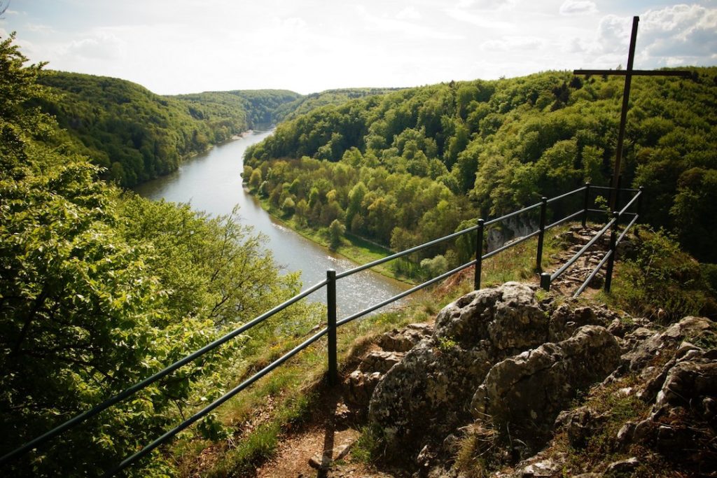 Das Bild ist an dem Aussichtspunkt „Wieser Kreuz“ oberhalb der Donau aufgenommen. Über eine Geländer auf einer Felskuppe ist die Donau inmitten von Wäldern zu sehen.