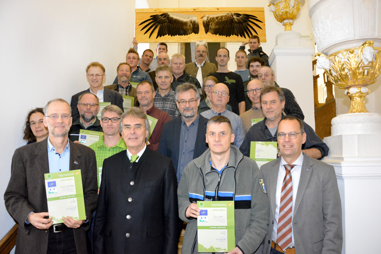 Eine Grupe überwiegend männlicher Personen steht auf einer Treppe. Im Hintergrund halten zwei Männer eine Tafel mit aufgedruckten Adlerflügeln hoch. Mehrere Personen halten Urkunden.