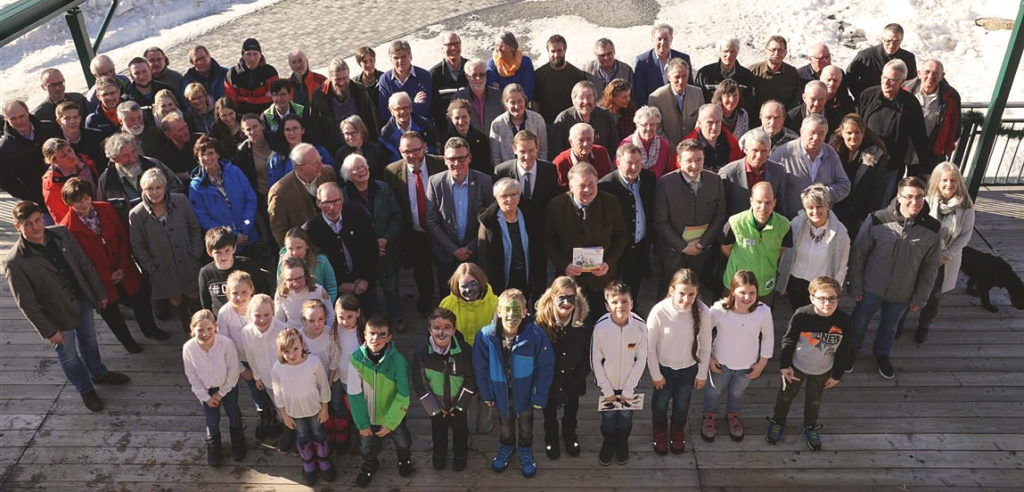 Eine große Gruppe Personen unterschiedlichen Alters steht im Freien vor einem Gebäude und blickt in Richtung des Fotografen nach oben.