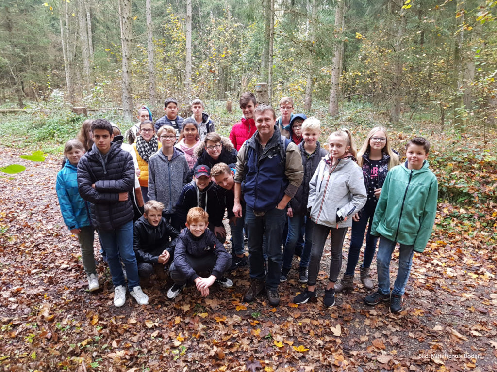 Eine Schulklasse mit Lehrer steht in einem herbstlichen Wald.