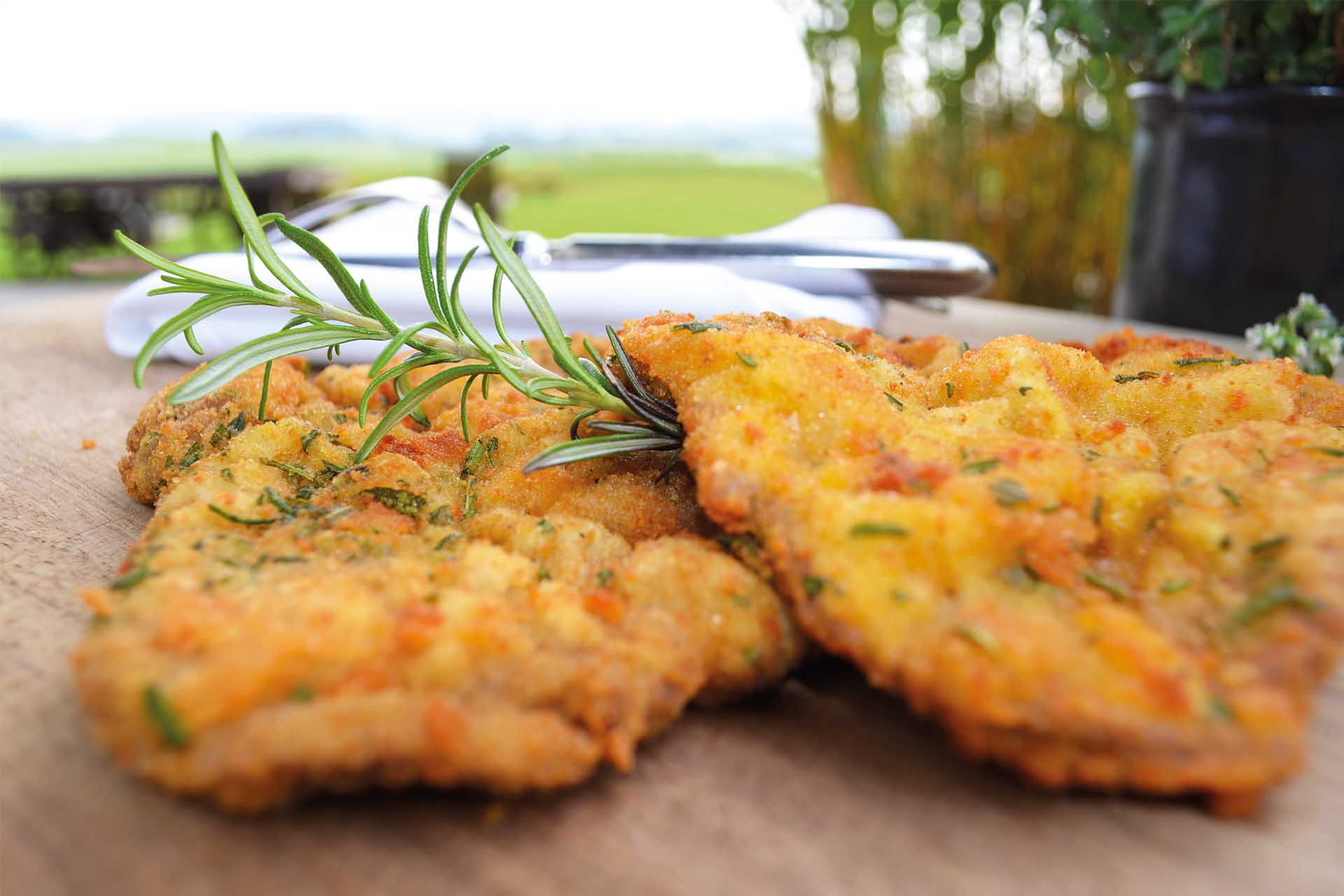 Zwei panierte und gebratene Schnitzel mit einem Rosmarinzweig auf einer Holzplatte.