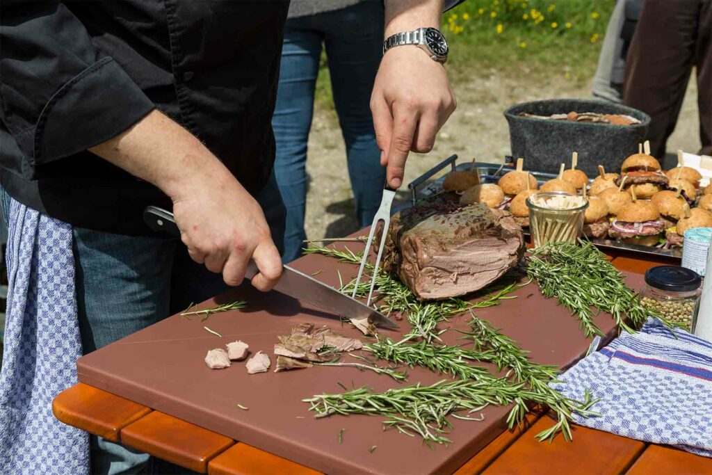 Lammbraten wird auf einem Schneidebrett mit einem großen Messer in kleine Stücke geschnitten, daneben liegen Kräuter.