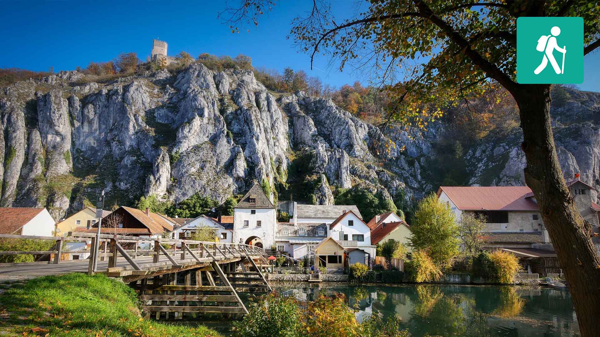 Eine Wiese mit einem Fluss und einer Holzbrücke, im Hintergrund ein Dorf und Berge.