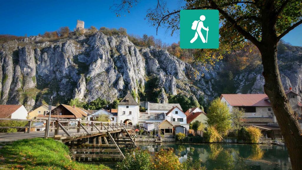 Eine Wiese mit einem Fluss und einer Holzbrücke, im Hintergrund ein Dorf und Berge.