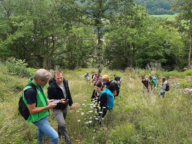 Eine Exkursionsgruppe steht auf einer Hangwiese und betrachtet Pflanzen