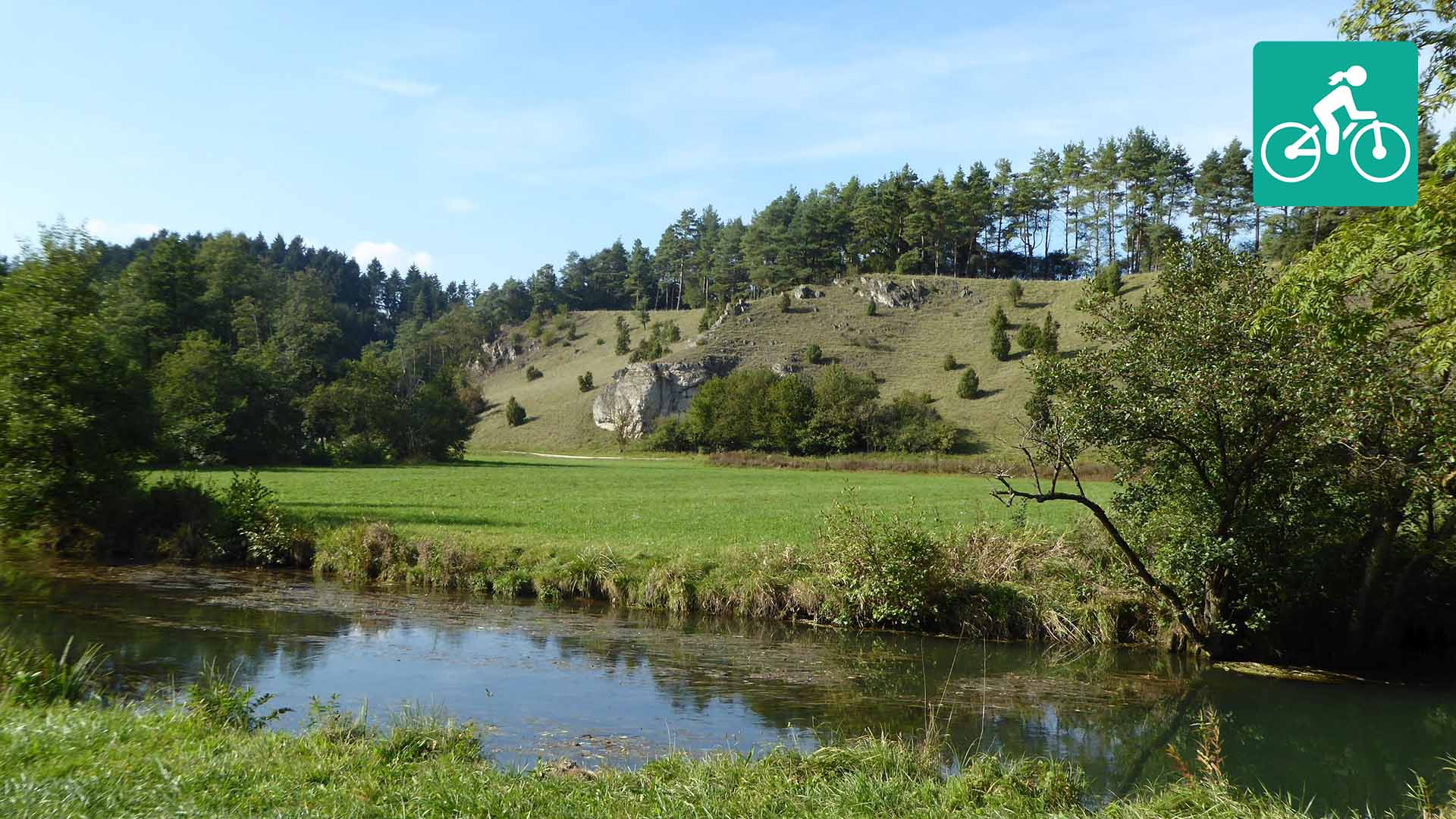 Ein Fluss fließt durch eine Wiese. Im Hintergrund ist ein bewaldeter Hügel zu sehen.