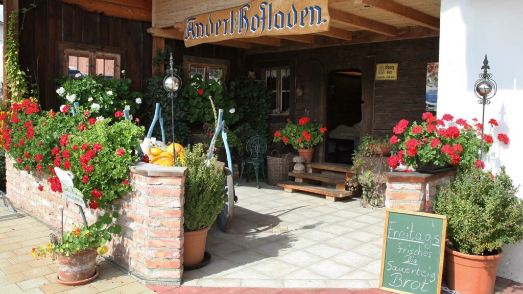 Eingangsbereich des Anderl Hofladen. Um das Holzhaus stehen viele Blumenkästen, sowie eine Holzkasten mit Gemüse.