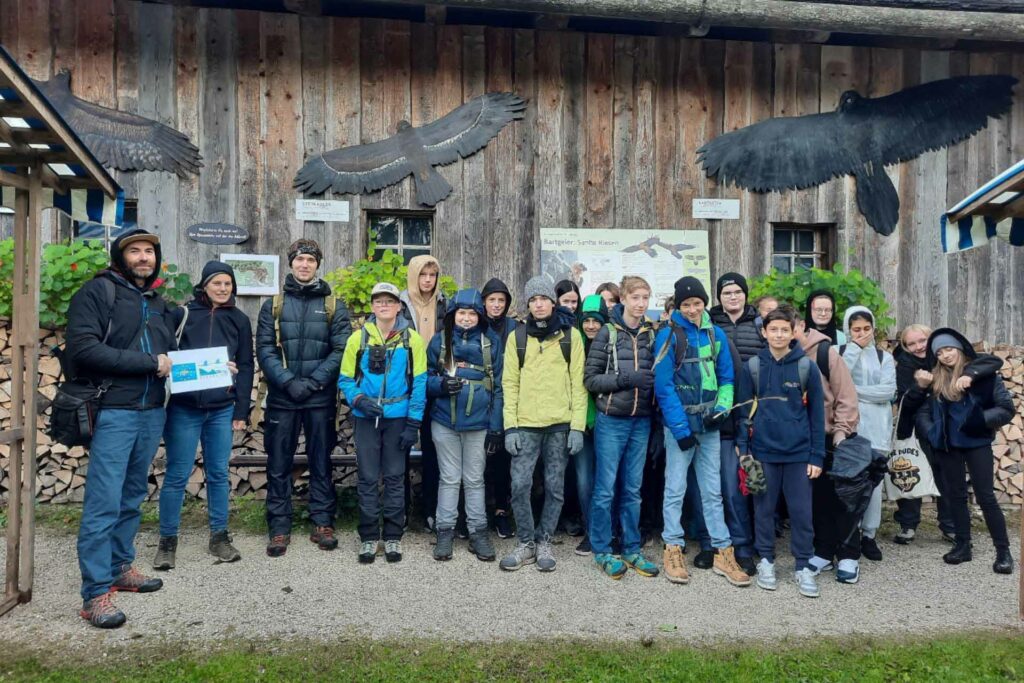 Eine Gruppe Jugendlicher steht vor einem Holzhaus. Ein Mann hält die Logos von LIFE und Natura 2000.