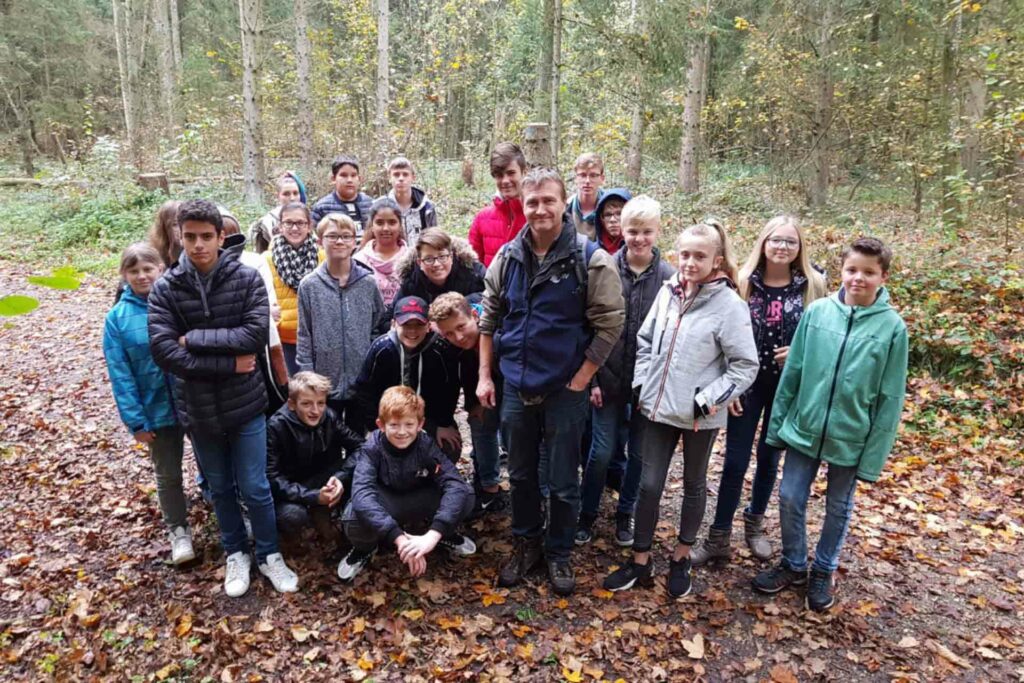 Eine Gruppe von Kindern steht mit einem Mann vor einem Wald.