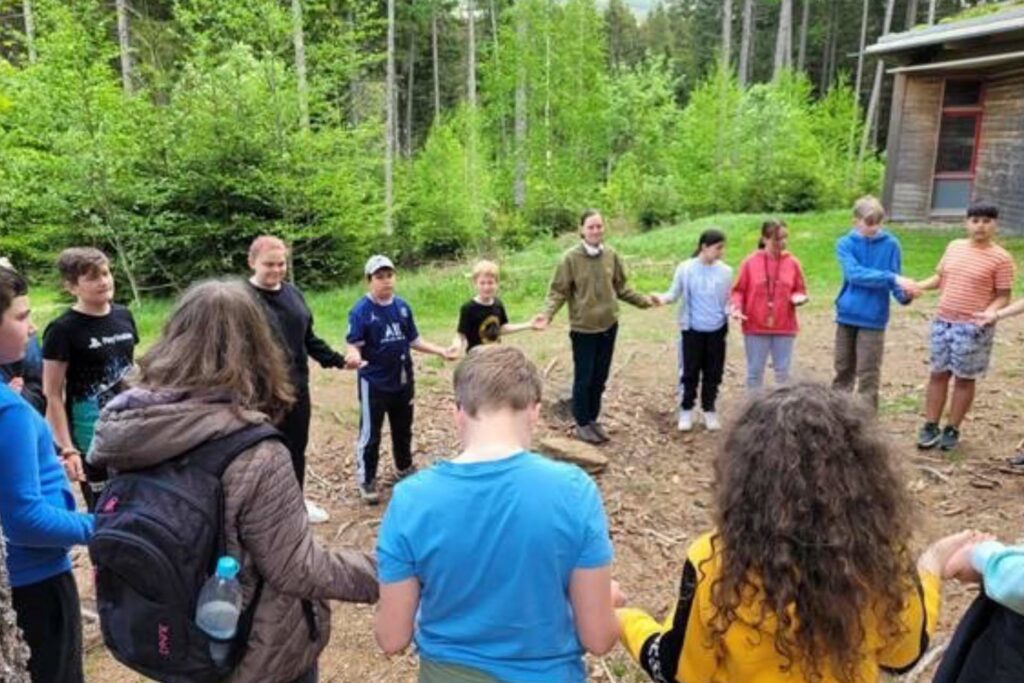 Schülerinnen und Schüler stehen in einem Kreis und halten sich an den Händen, im Hintergrund ist ein Wald und rechts ein Holzgebäude.