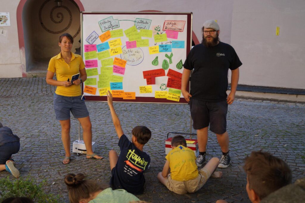 Eine Frau und ein Mann stehen vor einer Tafel mit bunten, beschriebenen Zetteln darauf. Davor sitzen Kinder am gepflasterten Boden.