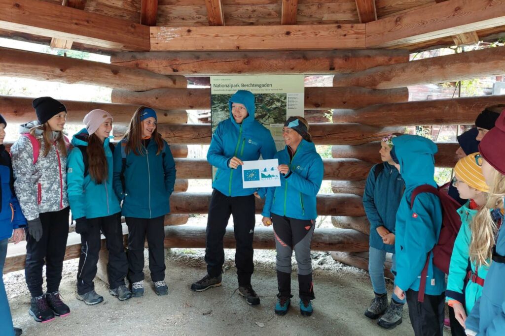 Zwei Personen stehen in einer Holzhütte und halten auf einem Zettel das Logo von LIFE und Natura 2000. Schülerinnen und Schüler sehen zu ihnen hin.