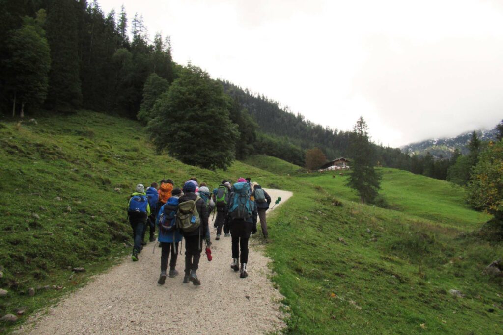 Eine Gruppe geht auf einem Weg durch eine Wiese. Im Hintergrund ist eine Almhütte zu erkennen.