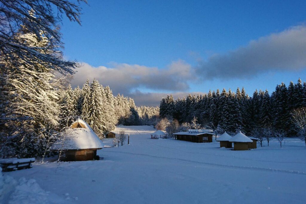 Holzhütten mit rundem Dach stehen auf einer offenen Fläche mit Nadelwald im Hintergrund. Alles ist schneebedeckt.