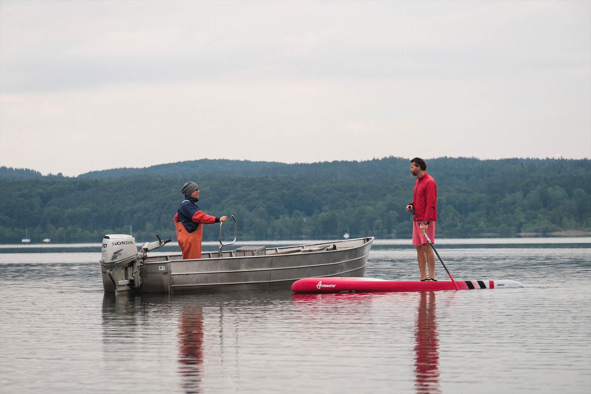 Blick auf den Starnberger See. Auf dem See treffen sich eine Fischerin und ein Stand-up-Paddler. Beide begrüßen sich freundlich.