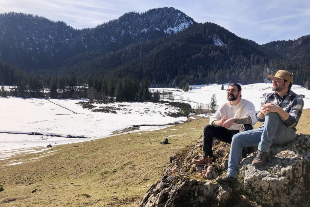 Flo Starflinger und Kamil Müller sitzen auf einem großen Stein bei Sonnenschein. Im Hintergrund sieht man ein weitläufiges Tal umringt von Bergen.