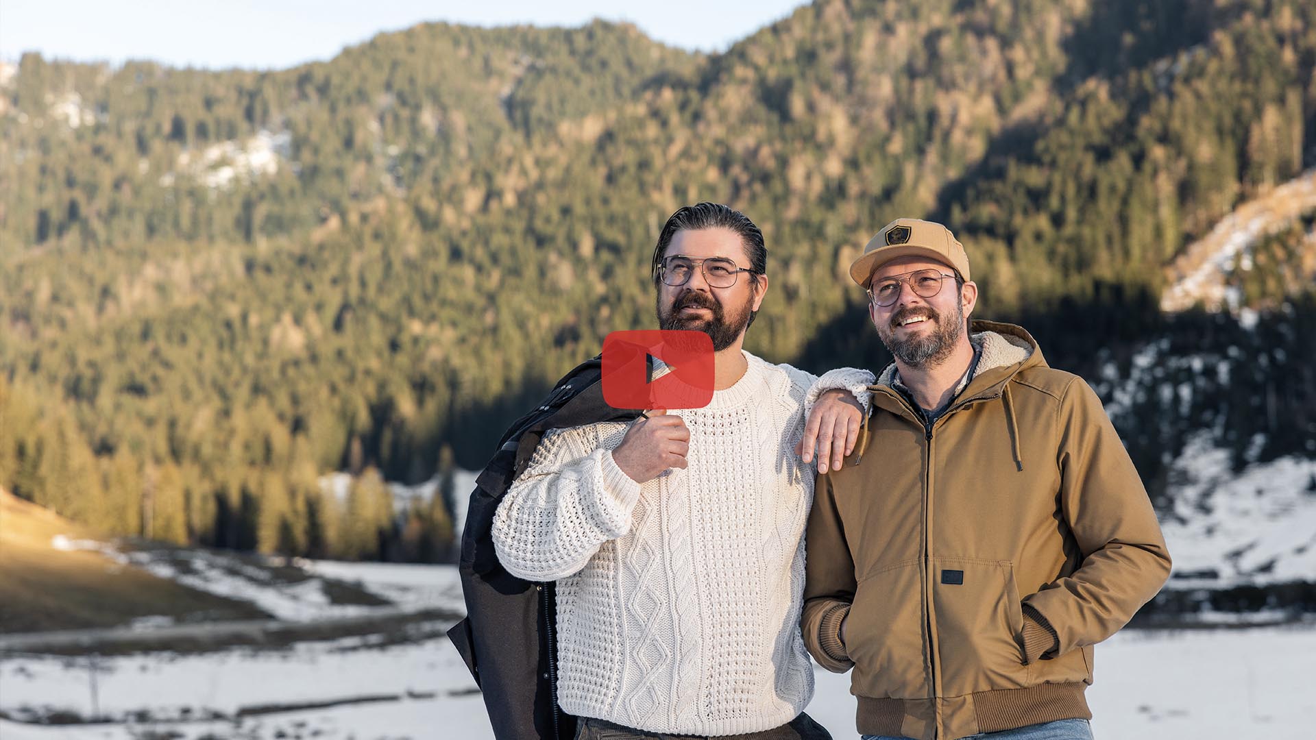 Florian Starflinger und Kamil Müller stehen in einem Bergtal. Im Schatten liegt noch Schnee, aber die Bäume leuchten schon grün in der Sonne.