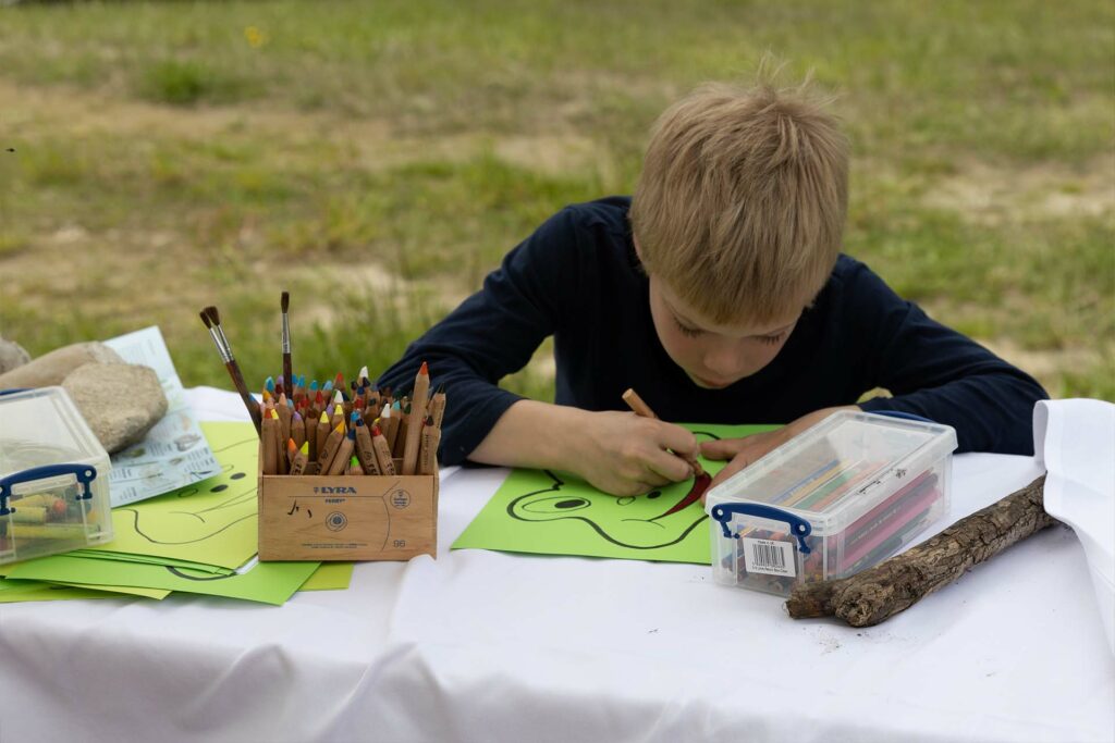 Ein Kind sitzt am Tisch und bemalt eine vorgedruckte Froschmaske.