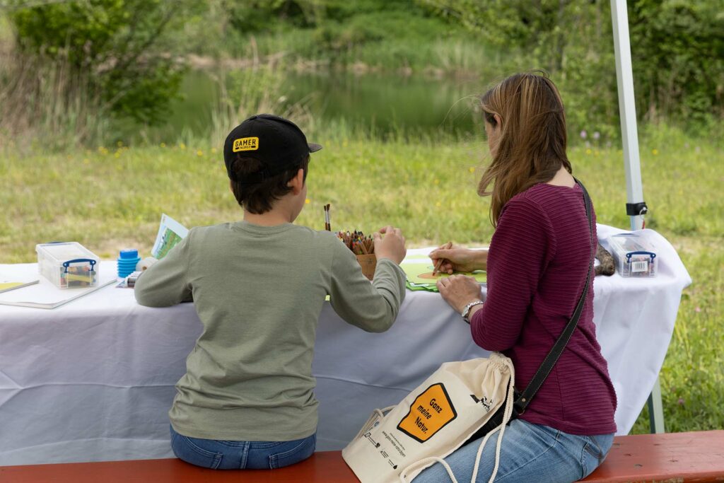 Eine Frau und ein Kind sitzen an einem Tisch mit Bastelmaterialen und basteln eine Froschmaske.